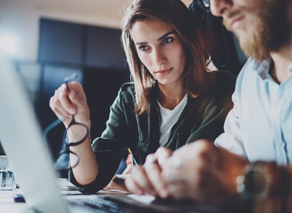 Woman holding her glasses while pointing out something on laptop screen to her colleague